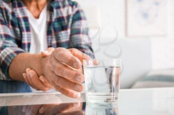 Senior man with Parkinson syndrome taking glass of water from table, closeup�