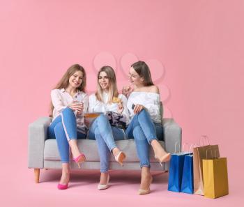 Young women with shopping bags, credit cards and mobile phone sitting on sofa against color background�