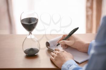 Woman writing something in notebook on table with hourglass in office. Time management concept�