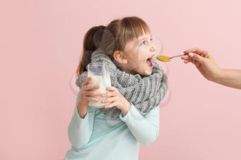 Little girl ill with flu holding glass of milk and eating honey on color background�