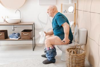 Elderly man with hemorrhoids sitting on toilet bowl in restroom�