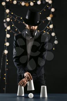 Magician showing tricks with cups on dark background�