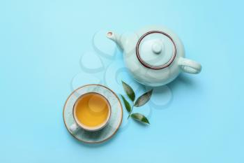 Teapot and cup of tea with green leaves on color background�