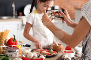 Female food photographer with mobile phone and little girl cooking pizza in kitchen�