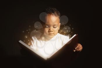 Little African-American boy reading magic book on dark background�