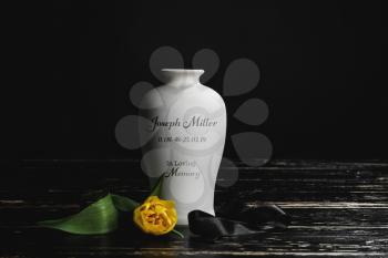 Mortuary urn with ribbon and flower on table against dark background�