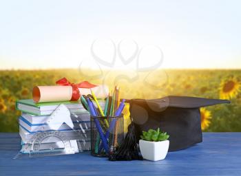 Mortar board, diploma, stationery and books on table near sunflower field�