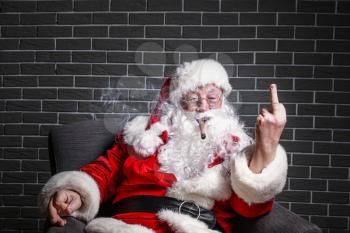 Funny drunk Santa Claus with cigar showing middle finger while sitting in armchair against brick background�