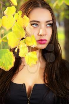 Young thoughtful brunette posing behind yellow leaves