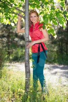 Smiling blond woman in red blouse and blue jeans posing near the tree