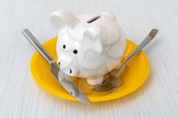Savings consumer concept. Close up view of piggy bank with a fork and knife on the plate 