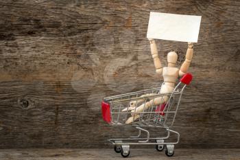 Wooden dummy sitting in a shopping cart and holding blank placard. Copy space.