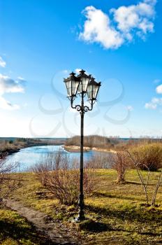 Black lantern with three lights on the background of blue sky, paths, grass, trees and blue of the river on a sunny day