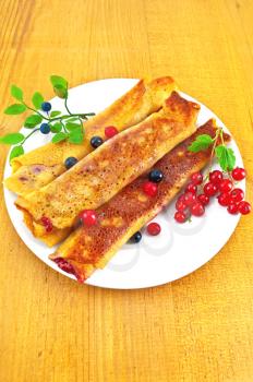 Pancakes with berry filling on a white porcelain plate, two brushes of red currant and blueberry twig with green leaves against a background of a wooden board