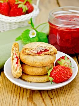 Stack of cookies filled with jam and strawberries on a plate, a jar of strawberry jam, a wicker basket with a strawberry on wooden board