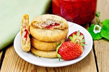 A stack of cookies filled with jam and strawberries on a plate, a jar of strawberry jam on the background of wooden boards
