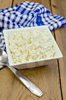 Cottage cheese in a white square bowl, a blue checkered napkin, spoon on a background of wooden boards