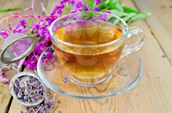 Herbal tea in a glass cup, a metal strainer with dry flowers fireweed, fresh flowers of fireweed on the background of wooden boards