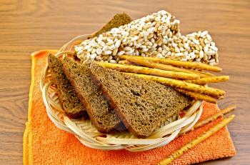 Fresh rye bread, bread sticks and cereal crispbreads in a wicker tray and the orange napkin against a wooden board