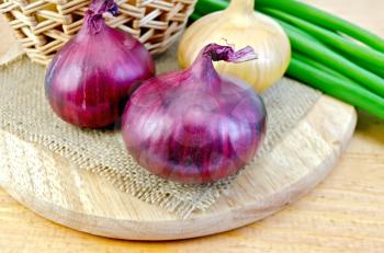 One yellow and two purple onions, a bunch of green onions, wicker basket on burlap background napkin and wooden boards