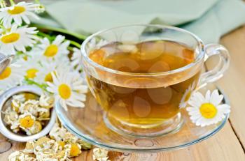 Herbal tea in a glass cup, metal sieve with dry chamomile flowers, fresh flowers, daisies, green cloth on a background of wooden boards