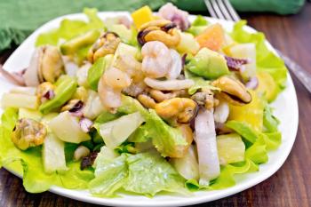Salad with shrimps, octopus, mussels and calamari with avocado, lettuce, pineapple in plate, napkin, fork on the background dark wooden boards