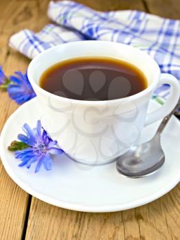 Chicory drink in a white cup with a spoon and a flower on a saucer, a blue checkered cloth on a wooden board