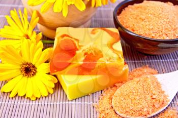 Two bars of homemade soap, bath salt orange, marigold flowers, a wooden mortar in the background of a bamboo napkin