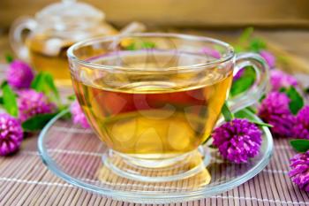 Herbal tea with flowers of clover in a glass cup and teapot on a bamboo napkin on a wooden boards background