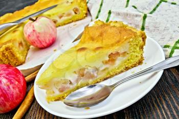 One slice of apple pie with cream sauce in white plate, spoon, cinnamon, apples and a napkin on a wooden boards background