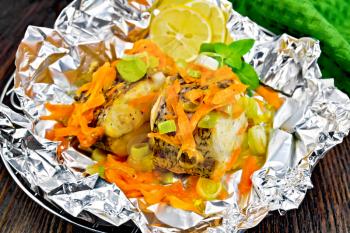 Pike with carrots, leek, basil and lemon slices on the grill in foil, a green towel on a wooden boards background