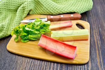 Rhubarb stalks with green leaves and a knife on a small planch, kitchen towel on a wooden boards background
