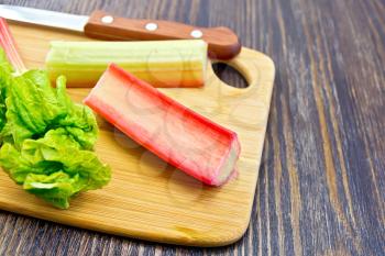 Rhubarb stalks with green leaves and a knife on a small planch on a wooden boards background
