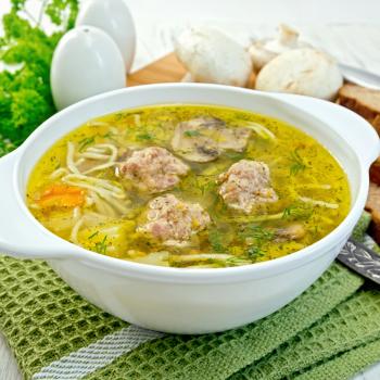 Soup with meatballs, noodles and mushrooms in a white bowl on a napkin, parsley on the background light wooden boards