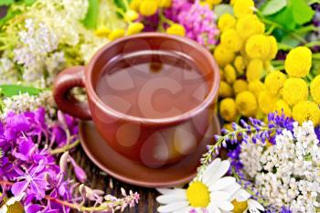 Herbal tea in a clay cup, fresh flowers fireweed, tansy, chamomile, clover, yarrow, meadowsweet, mint leaves on a wooden boards background