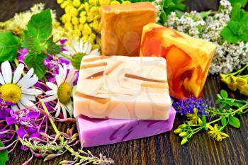 Four multi-colored bar of homemade soap, fireweed flowers, tansy, chamomile, clover, yarrow, tutsan and meadowsweet, mint and lemon balm leaves on the background of wooden boards