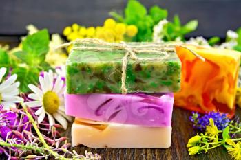 Three multi-colored bar of homemade soap, fireweed flowers, tansy, chamomile, clover, yarrow, tutsan and meadowsweet, mint and lemon balm leaves on the background of wooden boards
