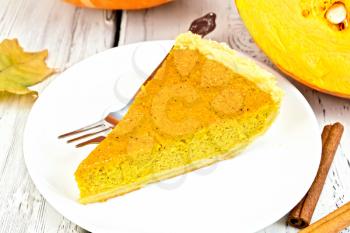 A piece of pumpkin pie with a fork in the white plate, cinnamon, pumpkin and maple leaf on the background light wooden boards