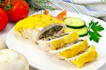 Roll of chicken breast with spinach, mushrooms and cheese in a white plate with slices of cucumber and tomato, napkin against the background of wooden boards