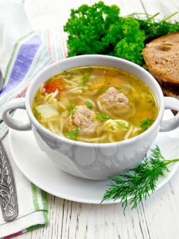 Soup with meatballs, noodles and vegetables in a white bowl and saucer, dill, bread, napkin and spoon on a wooden boards background
