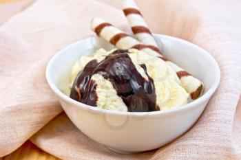 Vanilla ice cream in a white bowl with wafer rolls and chocolate syrup on a napkin against the background of wooden boards
