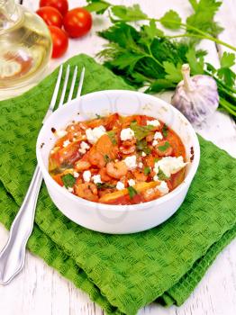 Shrimp and tomatoes baked with salty feta cheese in a white bowl on a green napkin, parsley and garlic, fork on the background of wooden boards