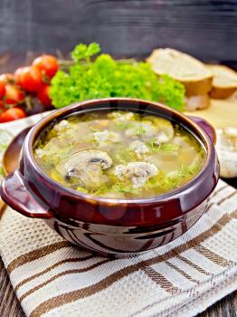Soup with meatballs, noodles and champignon in a clay bowl on a towel, parsley, tomatoes, mushrooms and bread on a wooden boards background