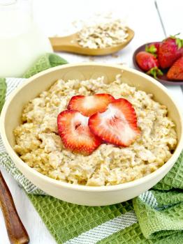 Oatmeal porridge in a bamboo bowl with strawberries on a green napkin, spoon, oat flakes in a spoon on a wooden board background
