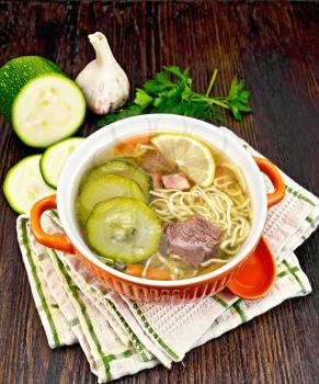Soup with zucchini, beef, ham, lemon and noodles in a bowl, parsley and dill on a napkin against a dark wooden board