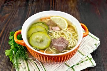Soup with zucchini, beef, ham, lemon and noodles in a red bowl, parsley and dill on a napkin on a wooden board background