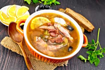 Soup saltwort with lemon, meat, pickles, tomato sauce and olives in a bowl on a sacking, bread on a background of a dark wooden board