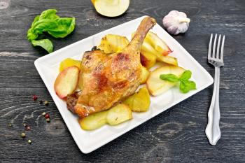 Roasted duck leg with apple, potatoes in a rectangular white plate, basil, garlic and fork on black background wooden plank