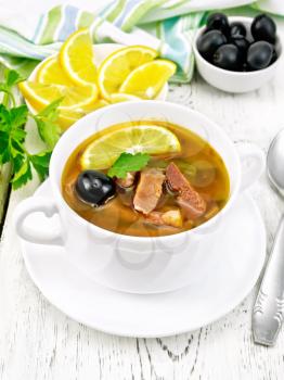 Soup saltwort with lemon, meat, pickles, tomato sauce and olives in a white bowl, towel, parsley on a wooden board background