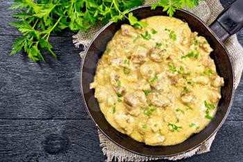 Meat stewed with cream in an old frying pan on burlap, parsley, fork on a wooden plank background on top